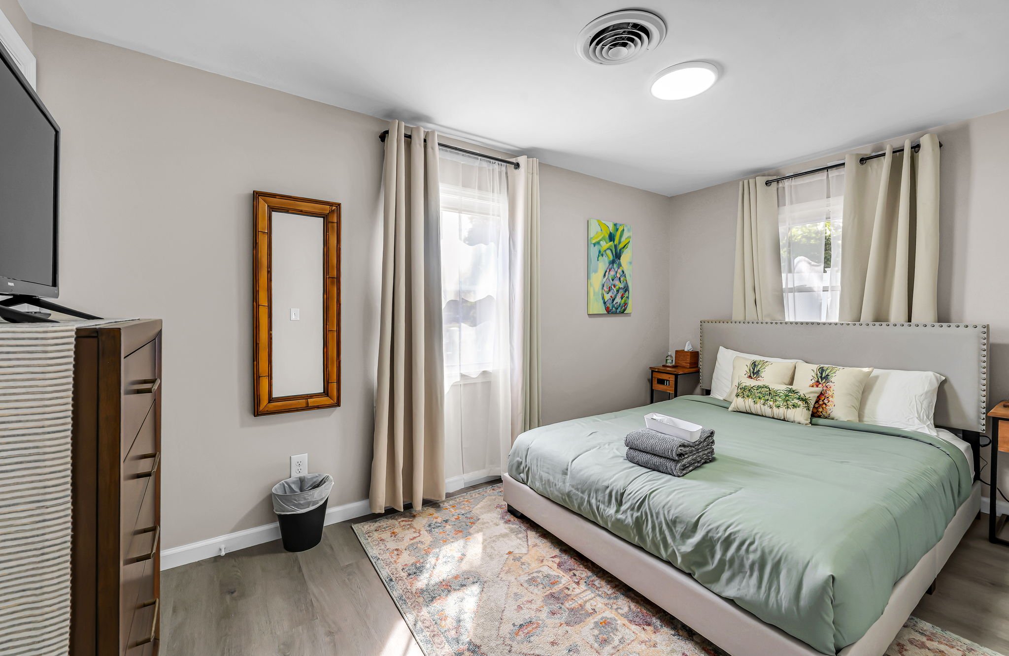 A bedroom with a bed, pillows, curtains, a window, a colorful painting, a mirror, a dresser, and a trash bin.