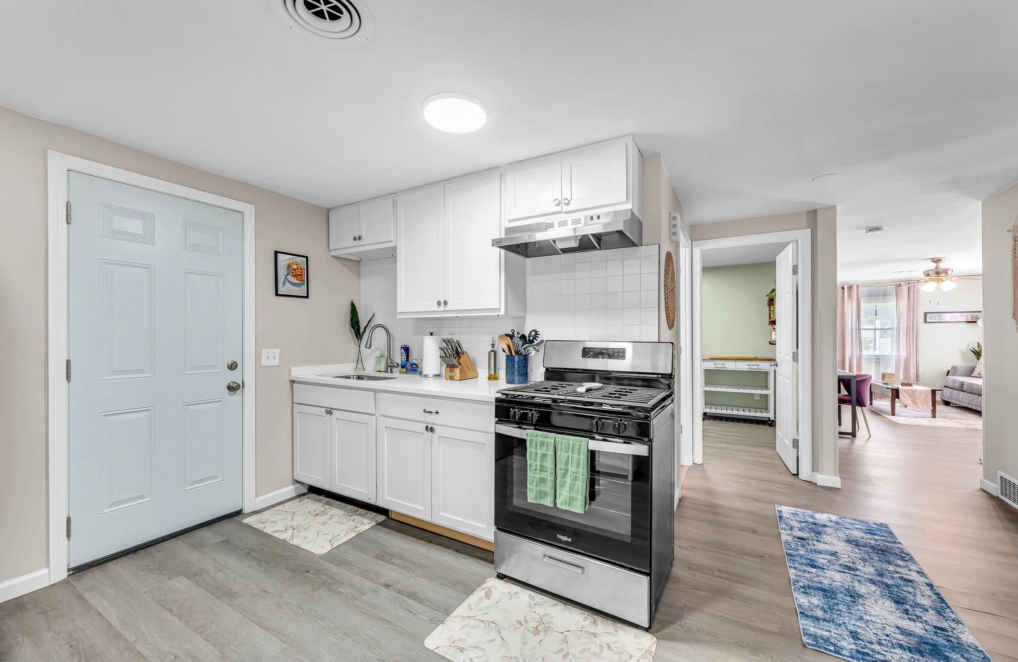 Modern kitchen with white cabinetry, stainless steel stove, gray wood flooring, and a doorway leading to a living room area.