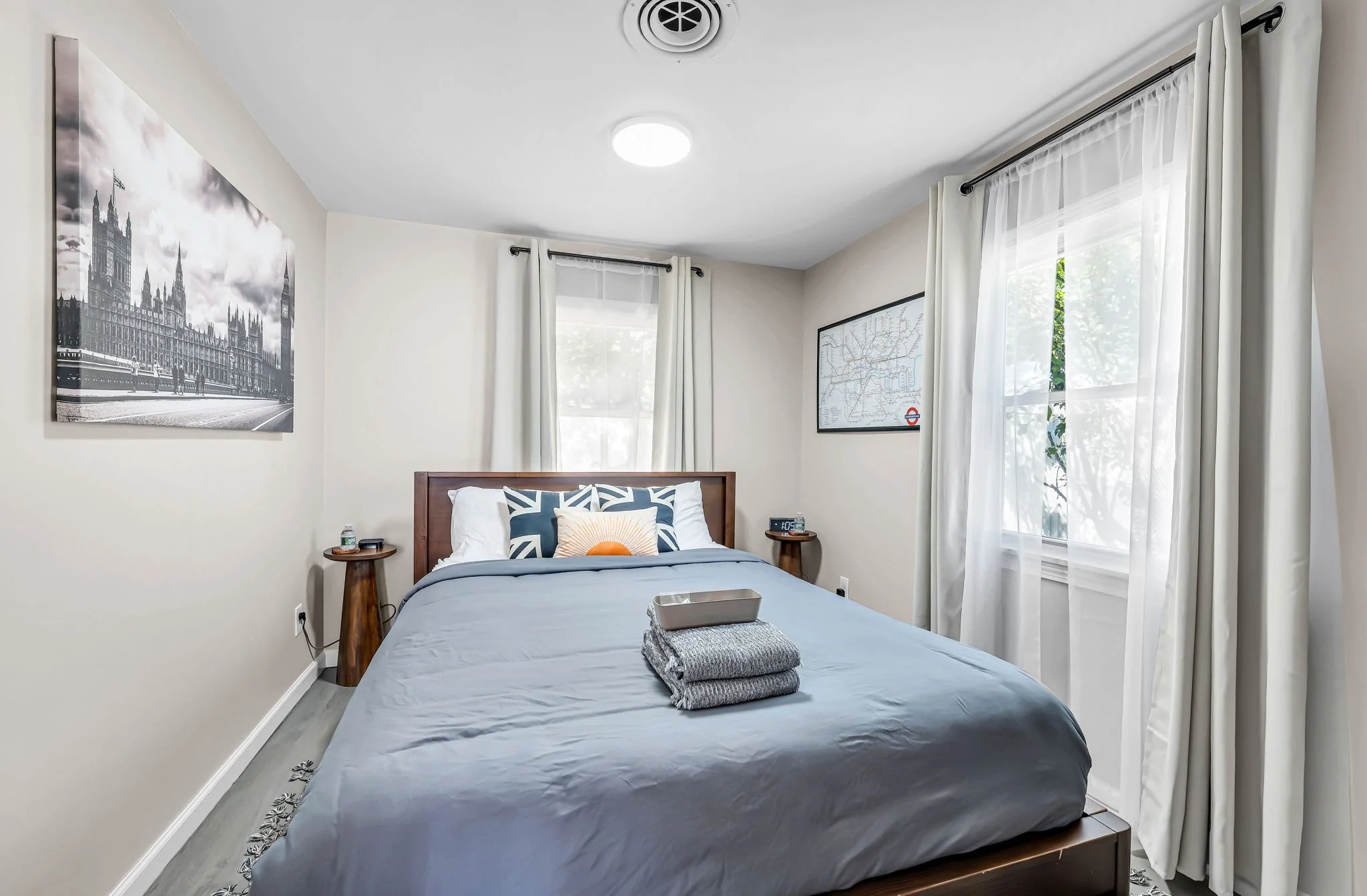 A bedroom with two windows with white curtains, a bed with a gray blanket and pillows, wooden nightstands with lamps, a black and white cityscape painting and a map of London on the walls, ceiling light, and a small tray and towels on the bed.