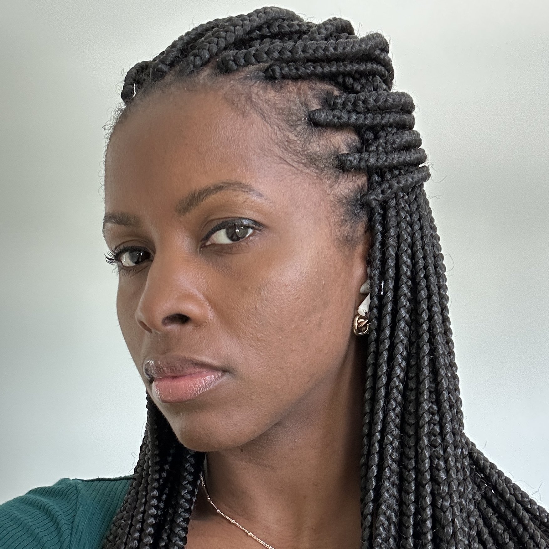 A woman with dark braided hair, wearing earrings, a green top, and a necklace, looking slightly to the side against a plain light-colored background.