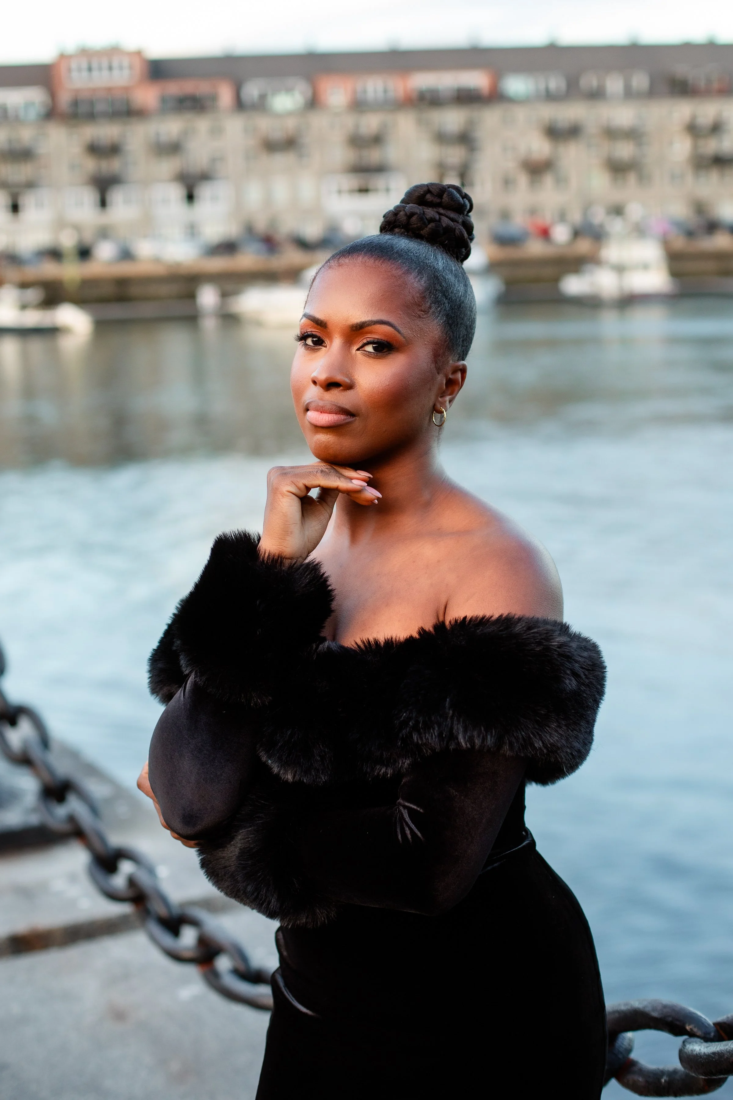 Portrait of a woman with dark hair styled in a braided bun, wearing a black outfit with fur trim, standing by a body of water with boats and a bridge in the background.