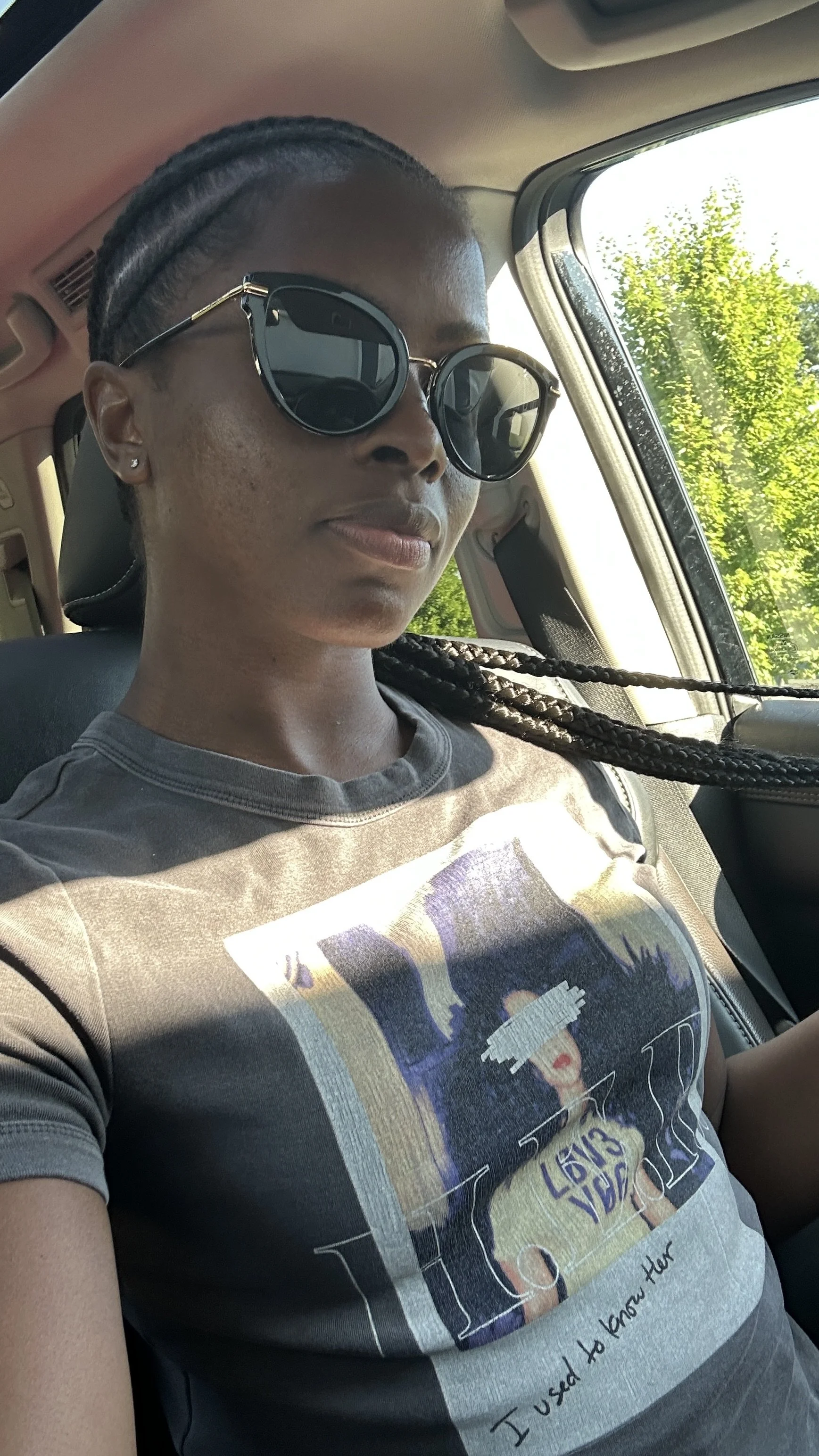 A woman wearing large black sunglasses and a graphic T-shirt sitting in a car with sunlight streaming through the window, with trees visible outside.
