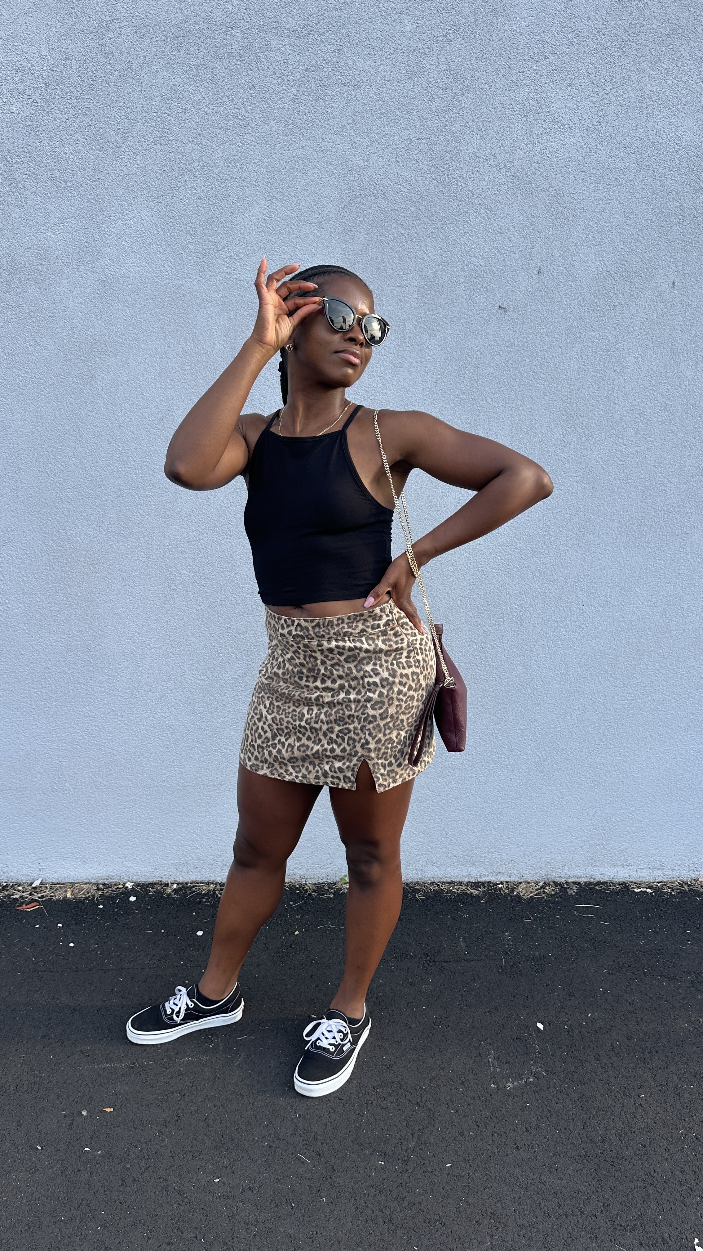 A woman stands against a plain textured light gray wall, wearing sunglasses, a black sleeveless crop top, a leopard print skirt with a small slit, and black sneakers with white soles. She has her left hand on her hip and her right hand adjusting her sunglasses, carrying a small burgundy shoulder bag.