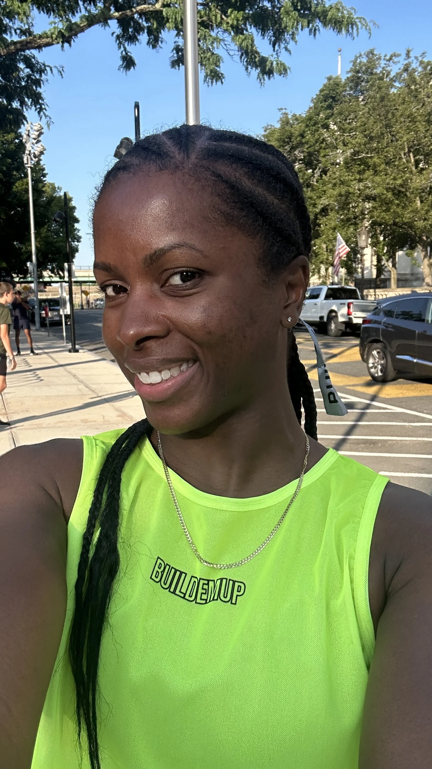 Close-up selfie of a smiling woman wearing a bright yellow athletic shirt with the text 'BUILD EM UP'. She has braided hair, earrings, a necklace, and appears to be outdoors in a parking lot with trees and cars in the background.