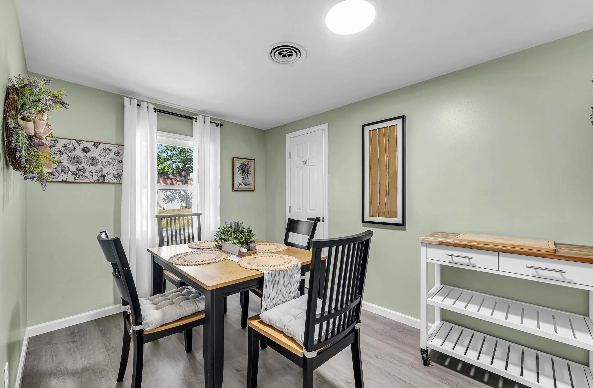 A cozy dining room with a wooden table and four black chairs with white cushions, a white sideboard, framed artwork on light green walls, and a window with white curtains letting in natural light.