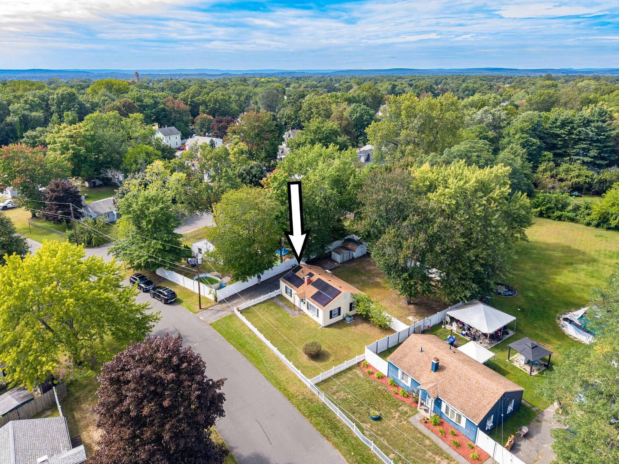 Aerial view of a suburban neighborhood with trees, houses, and a fenced backyard, with a distant horizon and partly cloudy sky.