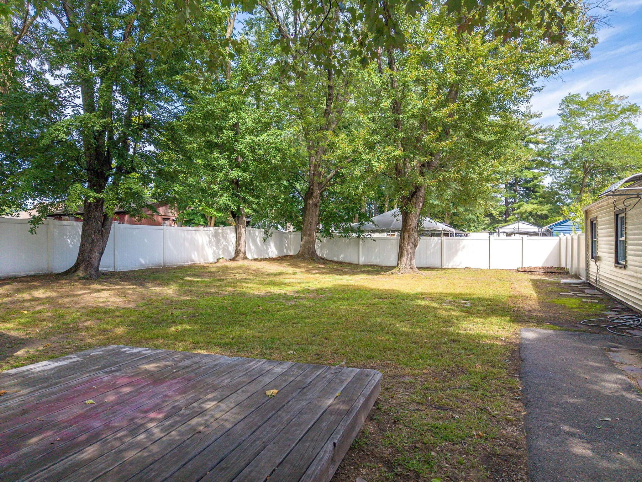 A backyard with a grassy area, surrounded by a white privacy fence. There are several large trees with green foliage. To the right, part of a house with siding and windows is visible, along with a garden hose on the ground. The sky is partly cloudy w