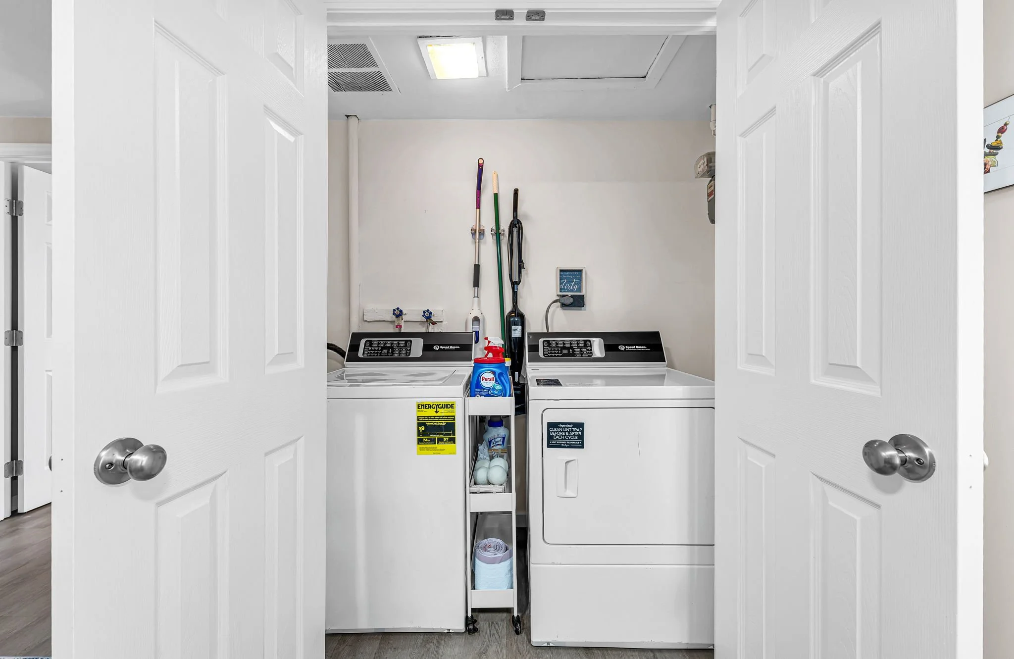 View inside a laundry closet with open double doors, showing a washing machine and dryer, cleaning supplies, and some cleaning tools on the wall.