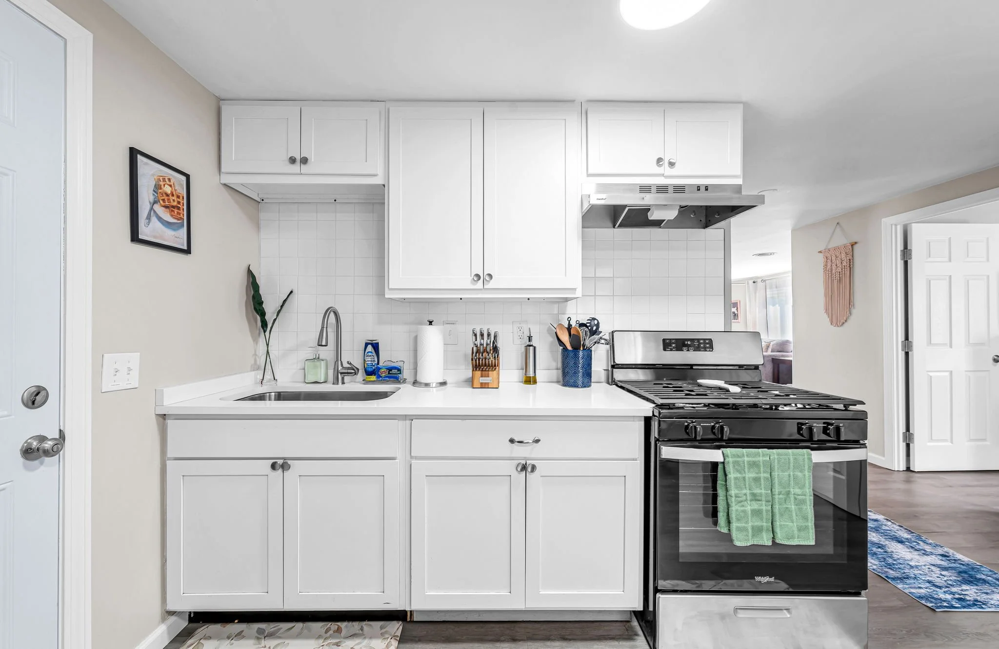 A modern kitchen with white cabinets, a black stove, and a white countertop. There are small kitchen tools and cleaning supplies on the counter, a green dish towel hanging from the stove, and a picture of waffles on the wall. The background shows a p