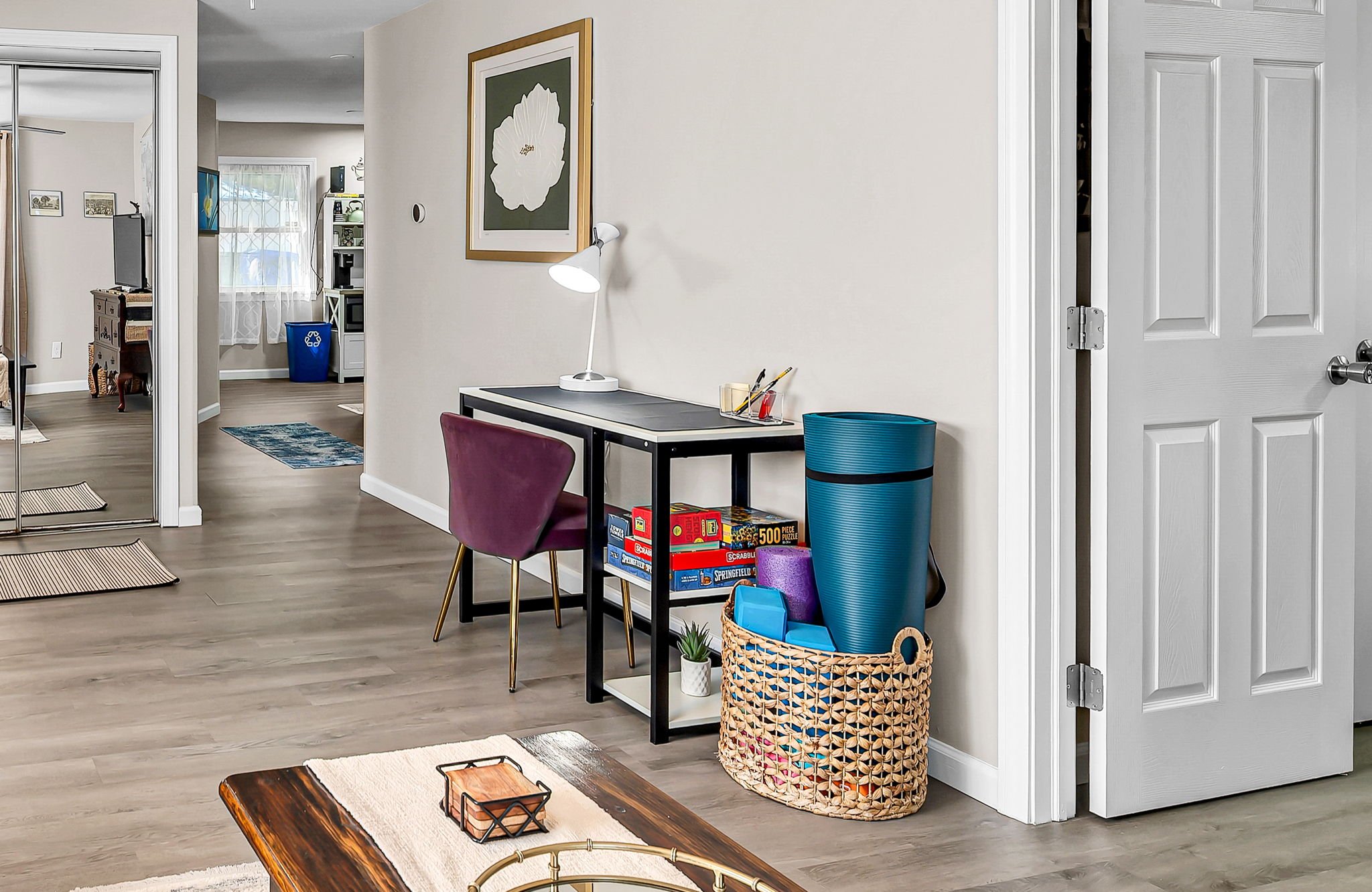 A room with a small black table, a maroon chair, and a desk lamp. There is a basket with a plant, a large blue trash bin, and a purple container. A linen on the coffee table has two candle holders. The room has wood flooring, and the walls are decora