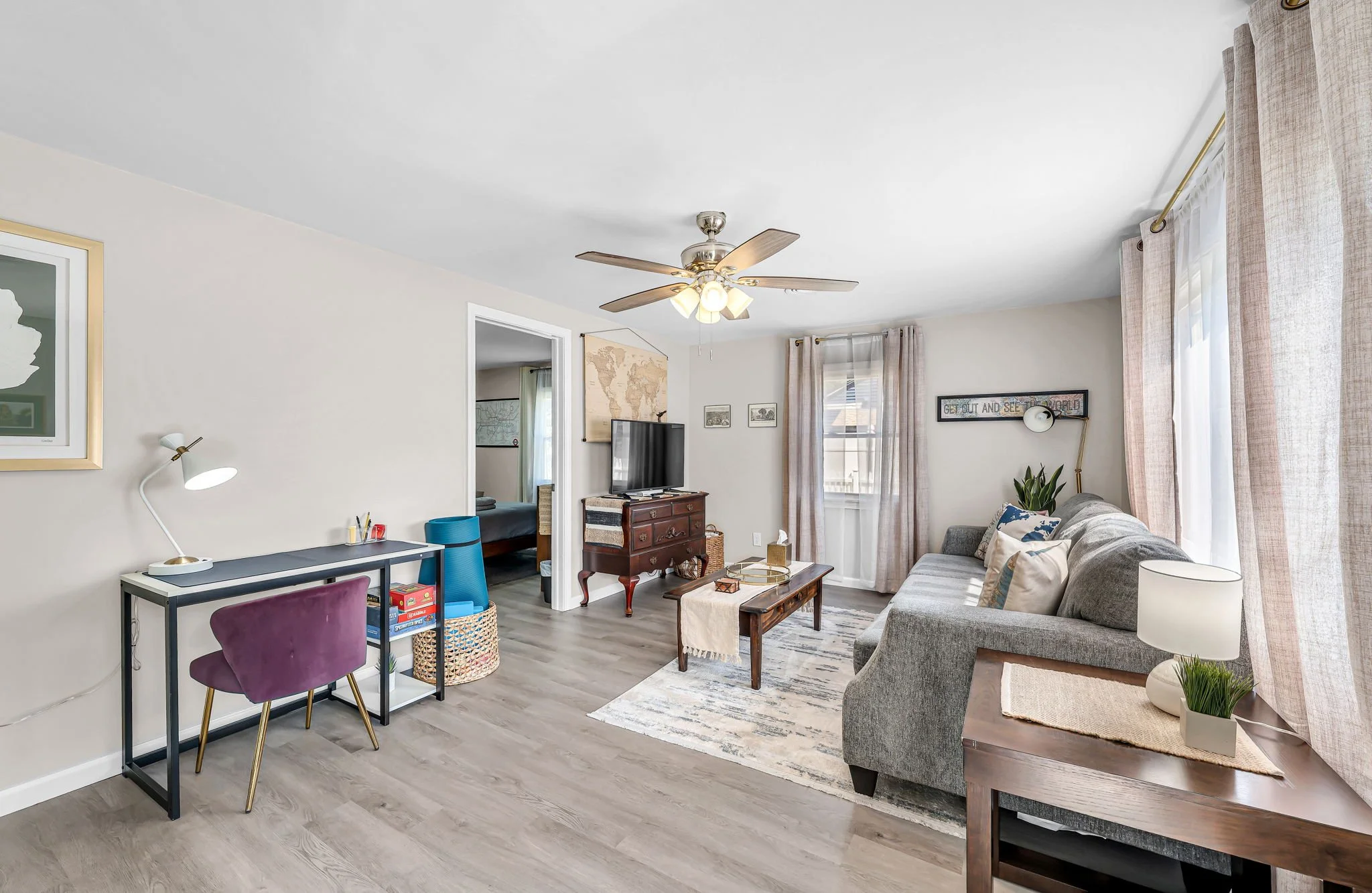 A living room with a ceiling fan, beige walls, and hardwood flooring. There is a gray sofa with pillows, a wooden coffee table, and a TV on top of a wooden dresser. The room has windows with beige curtains and a wall sign that says 'GET OUT AND SEE T