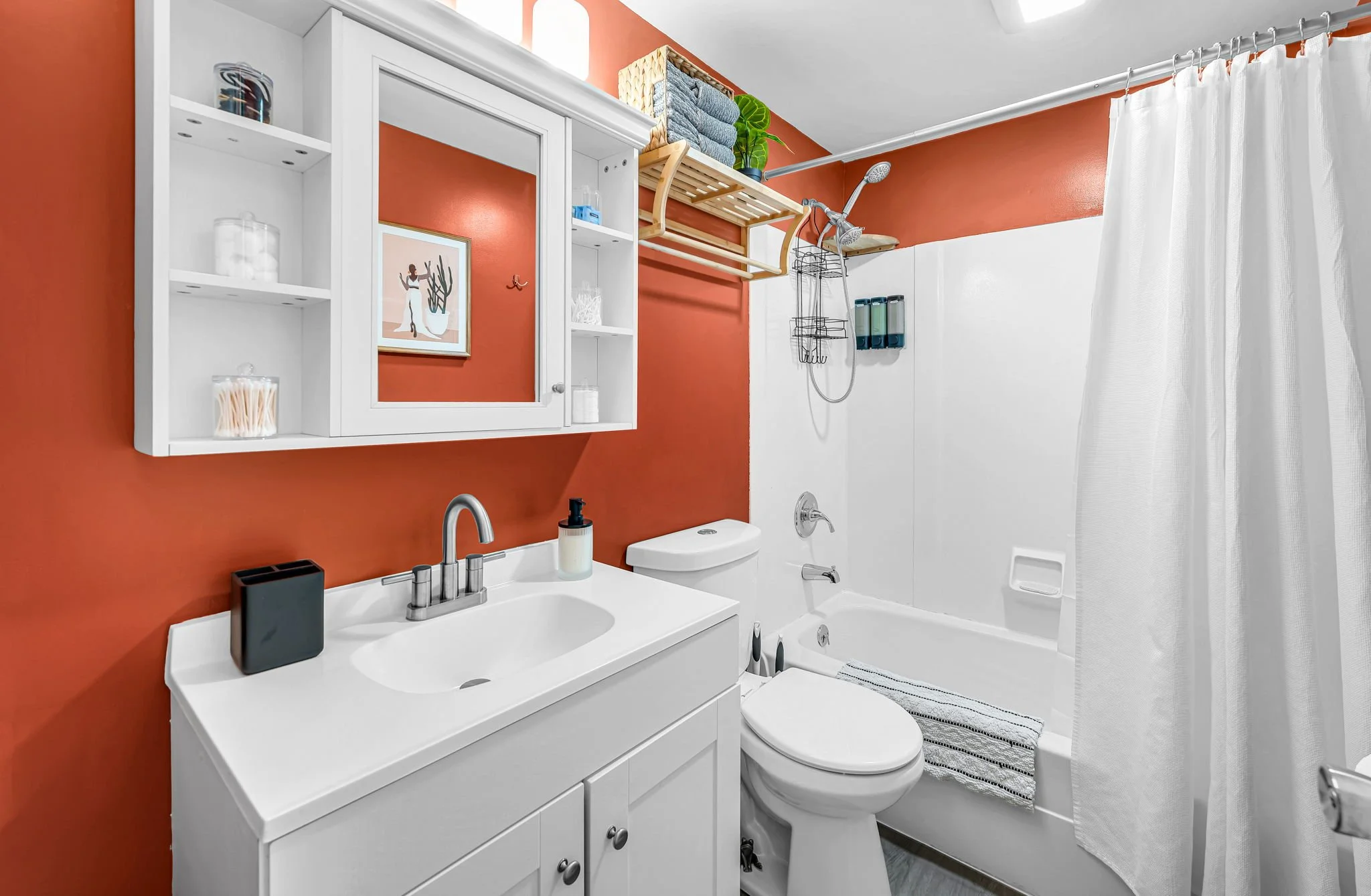 A bathroom with orange and white walls, featuring a white vanity with a sink, mirror, and toiletries, a toilet, a bathtub with a shower, and a white shower curtain. There are shelves and a towel rack above the bathtub, with folded towels and green pl