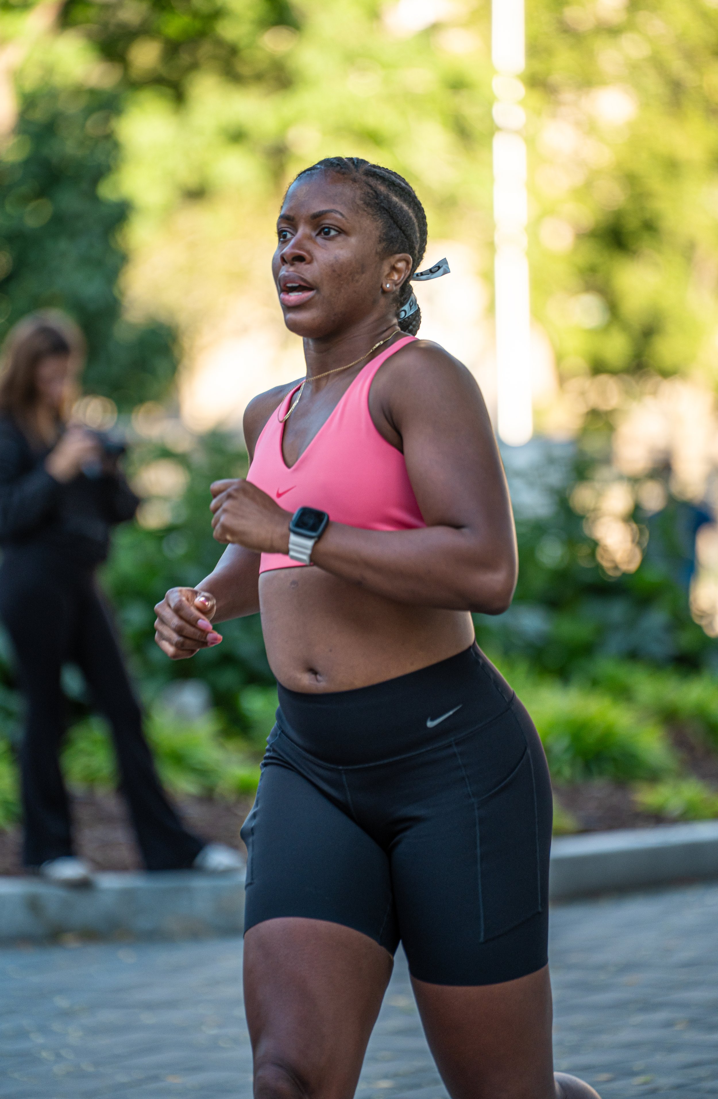 A woman running outdoors in a pink sports top and black shorts, wearing a smartwatch, with trees and a person in the background.