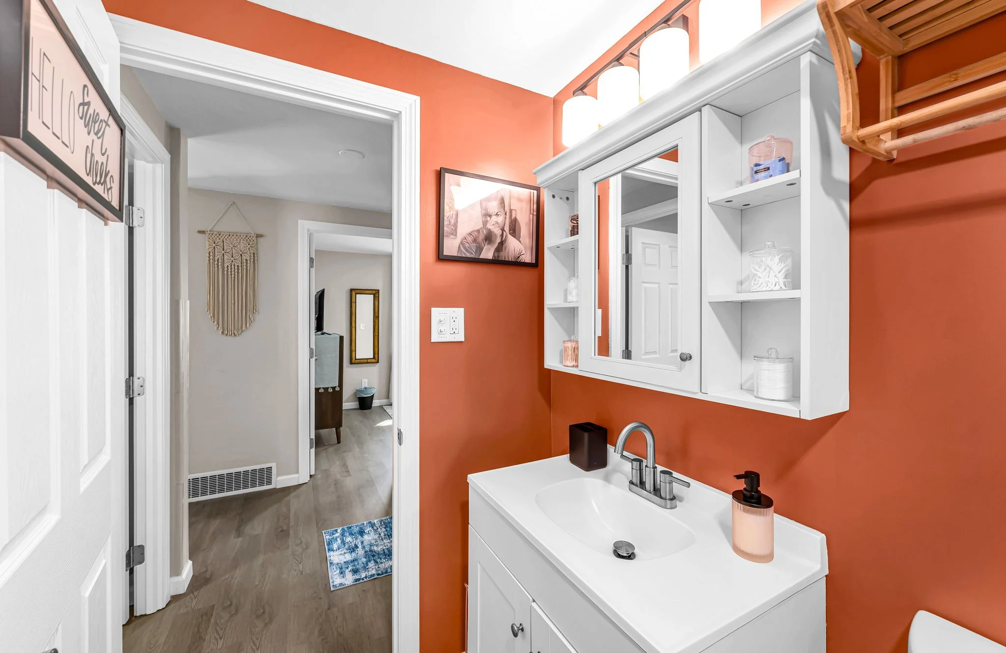 Interior view of a home with a white vanity and mirror above a sink on an orange wall, leading to a hallway with beige walls and decorative wall hangings.