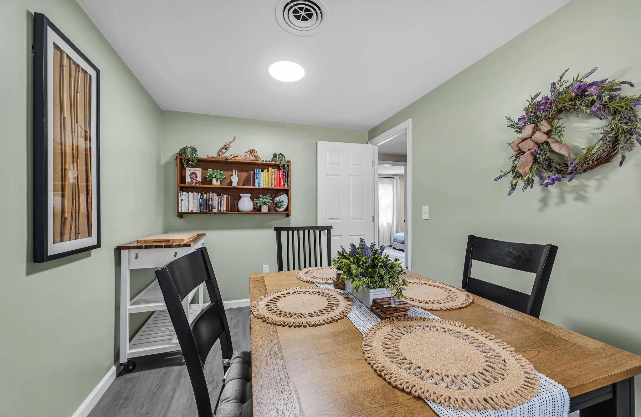 A dining room with light green walls, a wooden table with woven placemats and a floral centerpiece. There are black chairs around the table, a white bookshelf with books and decorative items, a framed artwork on the wall, a wreath with purple flowers