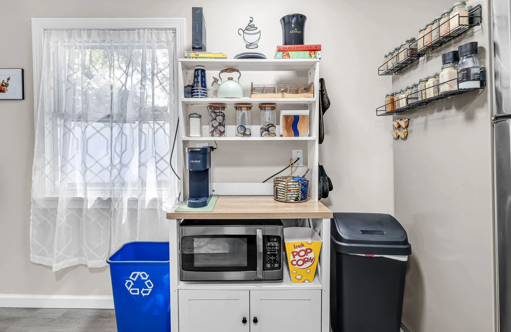 A kitchen corner with a white shelf and a mini oven, a microwave, and a popcorn box. There are jars of spices on a wall-mounted rack, a Keurig coffee maker, and a blue recycling bin. Light is coming through a window with a white curtain.