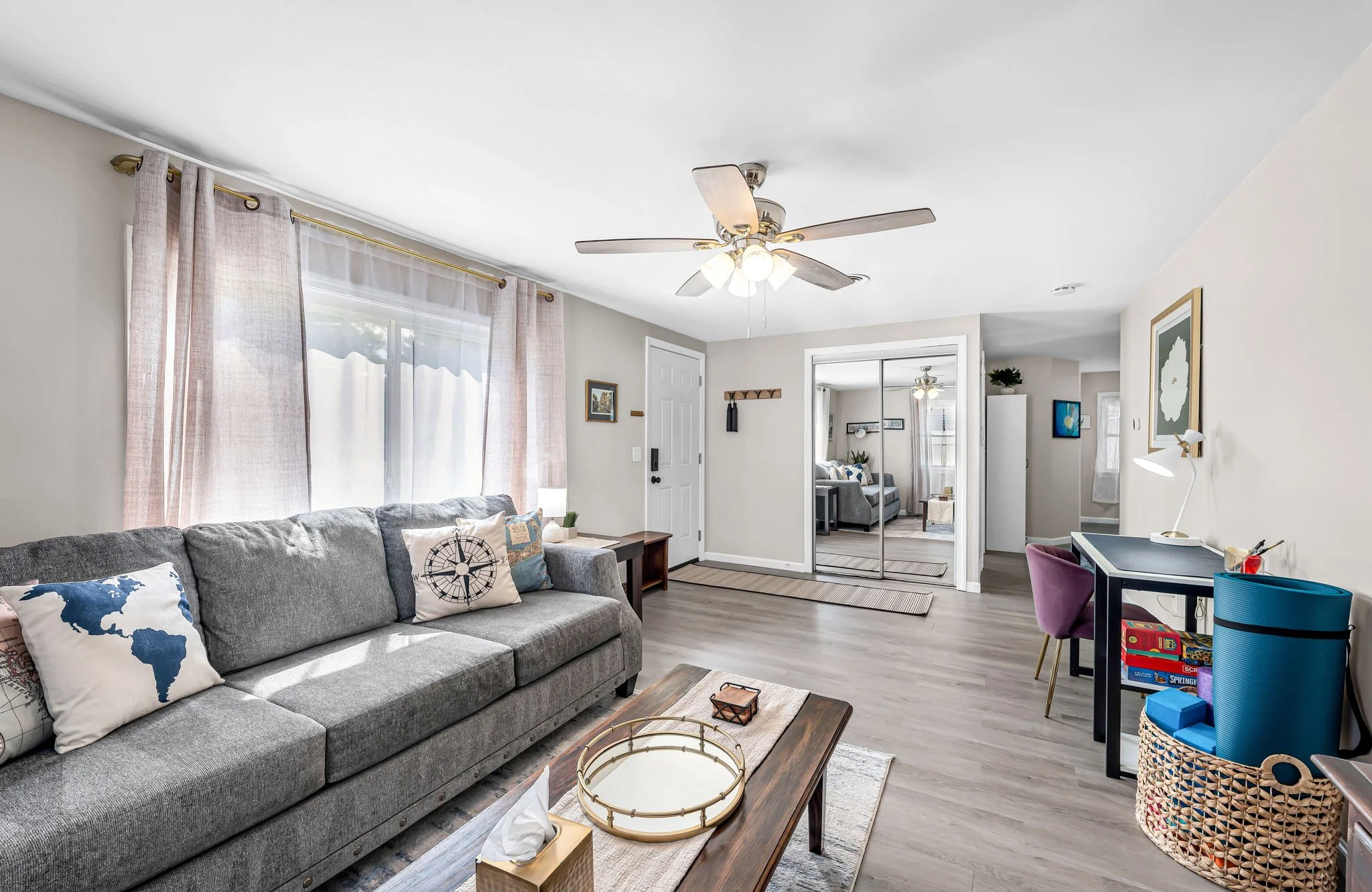 A living room with a gray sofa, decorative pillows, a wooden coffee table, and a ceiling fan with lights. There are windows with curtains, a mirror closet door, and various furniture and decorations.