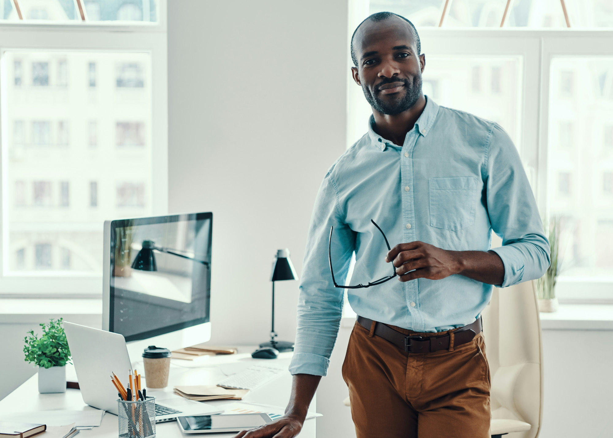 A man standing in an office, holding glasses, next to a desk with a computer, plant, coffee cup, and office supplies, with large windows in the background.