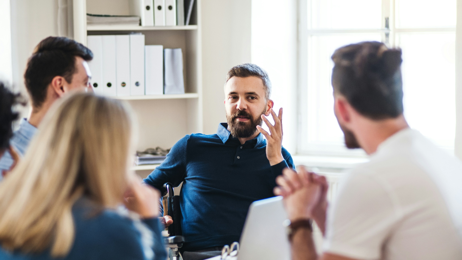A group of diverse people having a meeting or discussion in a bright room, with a man in a wheelchair speaking.