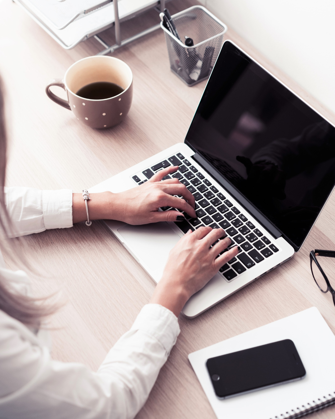 Person typing on a silver laptop at a wooden desk, with a cup of coffee, a smartphone on a notepad, eyeglasses, a pen holder, and a paper tray nearby.