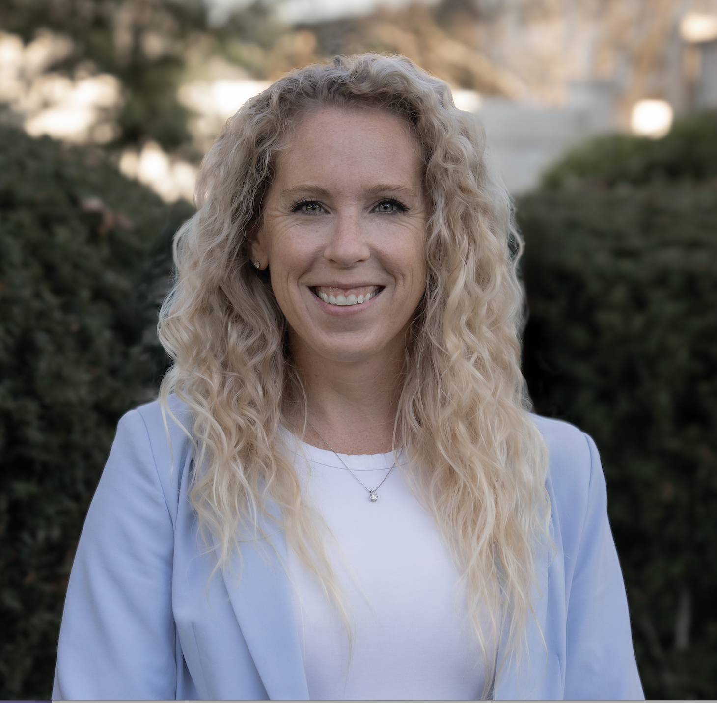 A woman with long, blonde, curly hair outdoors, smiling at the camera, wearing a light blue blazer and white top, with blurred greenery background.