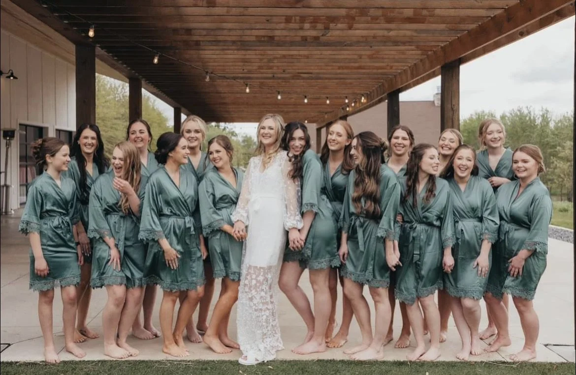 Bride in a white lace dress holding hands with a group of women in matching teal robes, all barefoot, standing under a wooden covered outdoor pavilion with string lights.