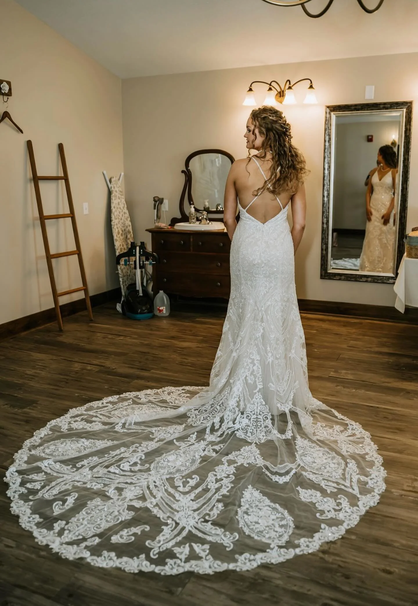 A bride in a white lace wedding dress with a long train standing in a bedroom, facing away from the camera. Her reflection is visible in a full-length mirror. The room has wooden flooring, an ironing board, a dresser with a mirror, and various items
