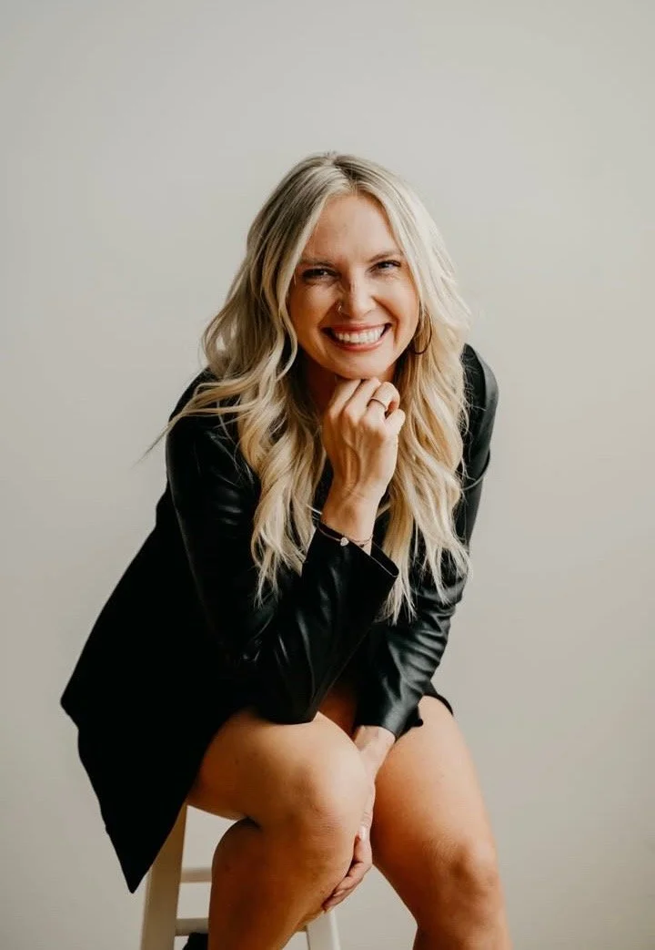 A woman with long blonde hair, smiling and looking at the camera, sitting on a white stool against a plain light-colored wall, wearing a black leather jacket and black shorts.