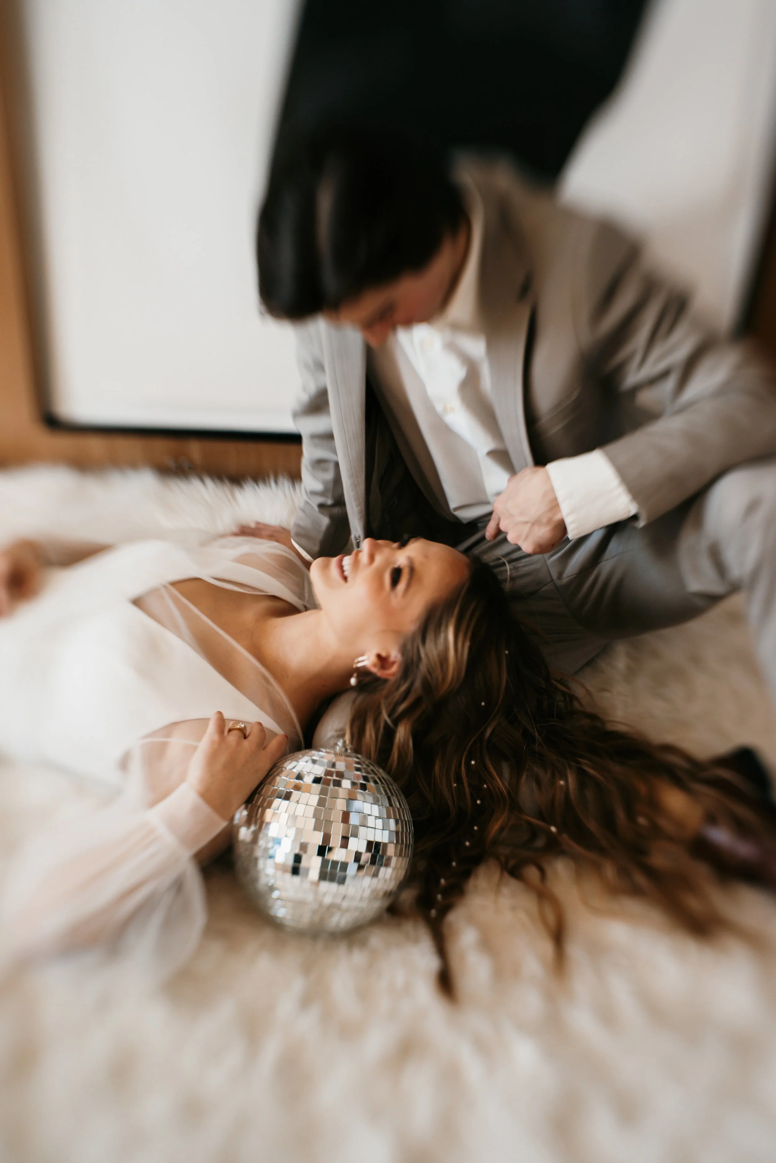 A woman lies on a fluffy surface with a disco ball near her head, smiling and looking up at a man in a suit, who is leaning over her.