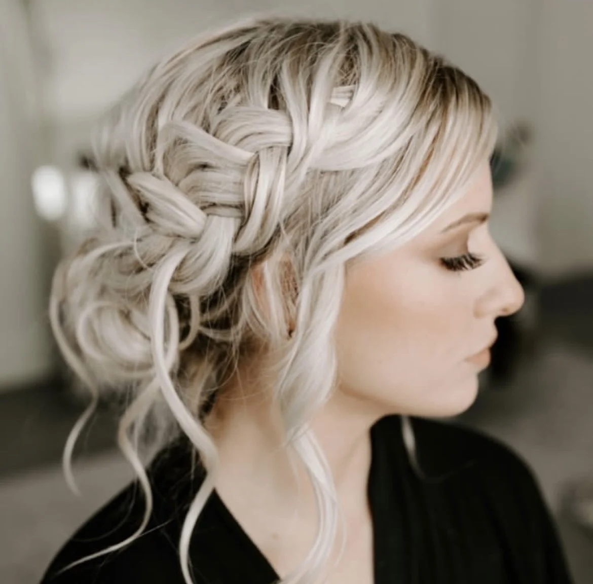 Close-up of a woman with platinum blonde hair styled in an intricate braid and loose curls, wearing a black top, in a softly lit room.