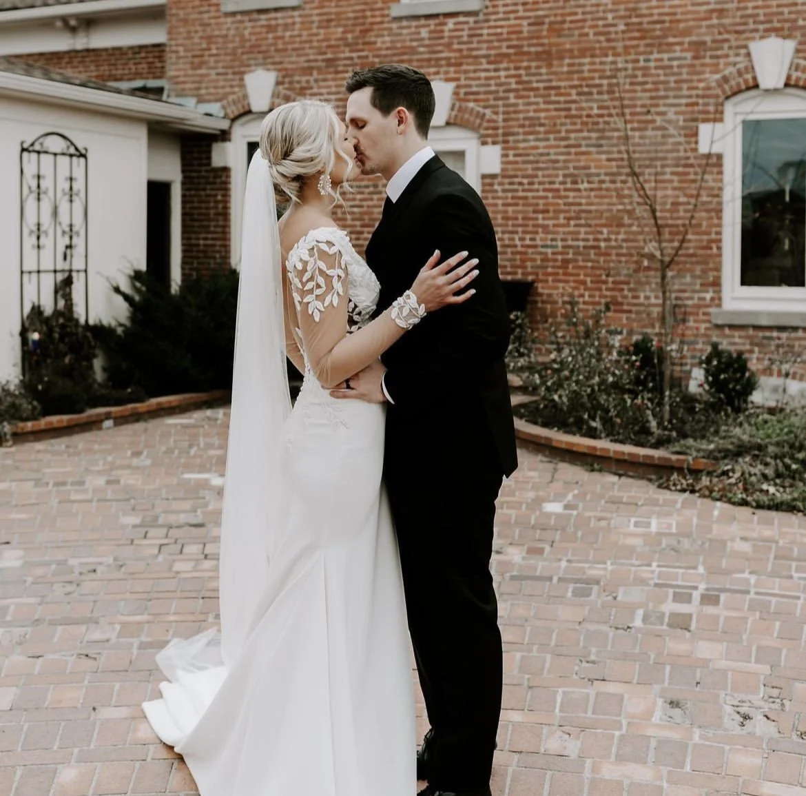 A bride and groom share a kiss outdoors on a brick patio, with a brick house and garden in the background.