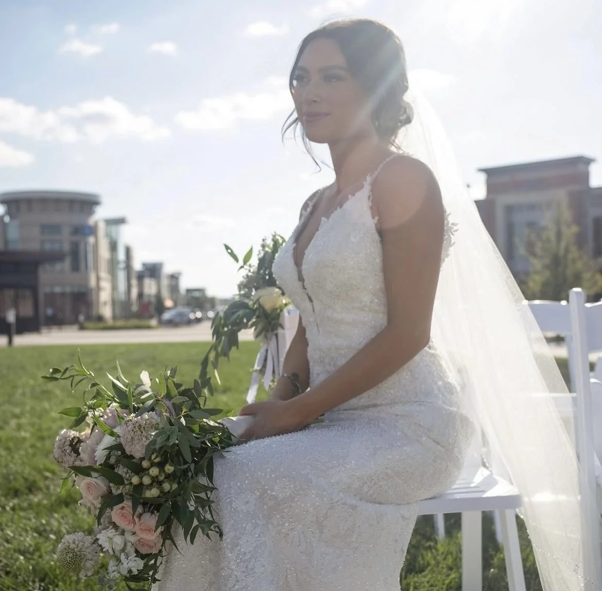 A bride in a white wedding dress sitting outdoors, holding a bouquet of flowers, with buildings and a blue sky in the background.