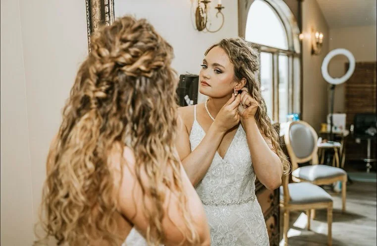 A bride with long, curly blonde hair gets ready, looking in the mirror as she puts on earrings in a well-lit room with elegant furniture.