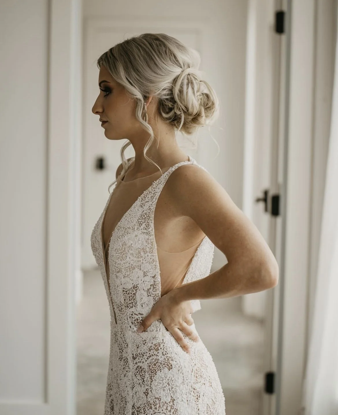 A woman in a wedding dress standing in profile with her hand on her hip in a room with light-colored walls and doors.