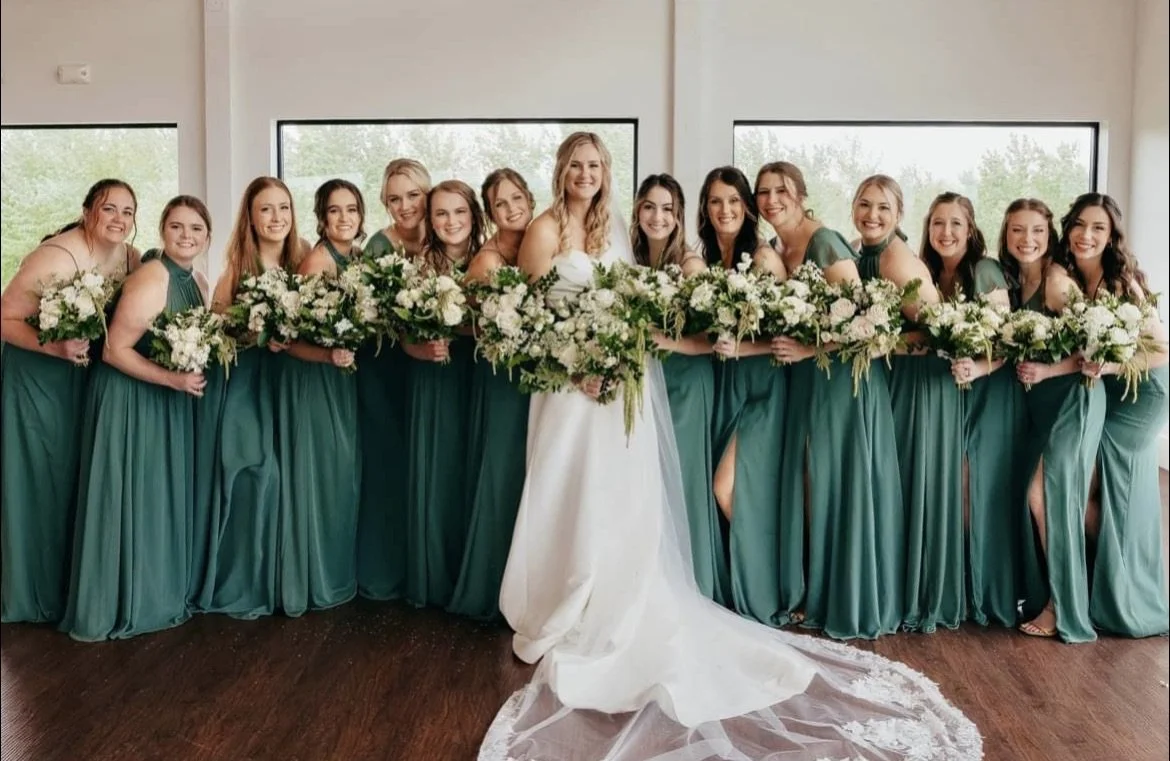 A bride and her 12 bridesmaids in teal dresses, holding bouquets, standing in a room with large windows