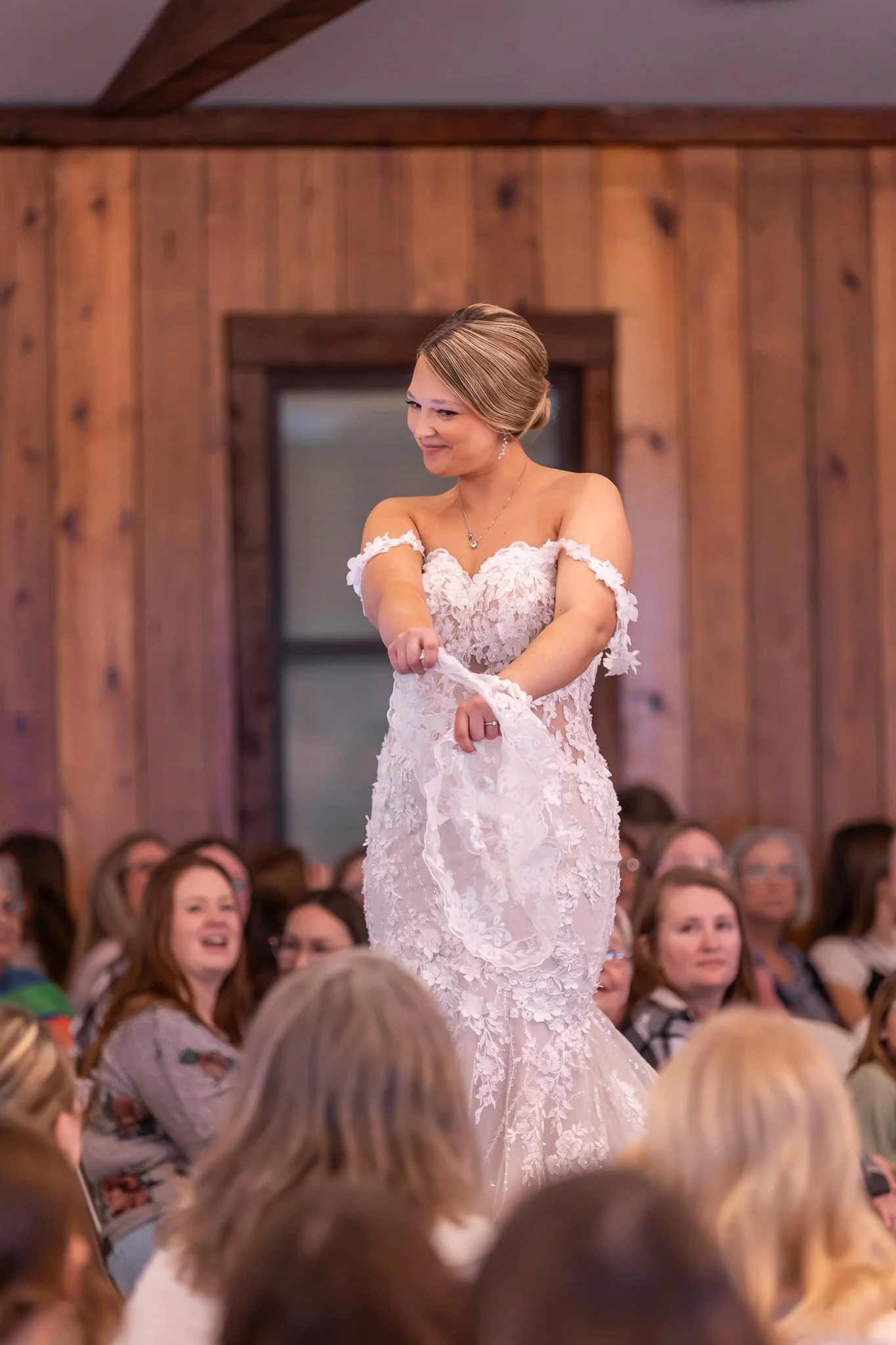 A bride in a white lace wedding dress with off-the-shoulder sleeves is smiling and adjusting her dress at her wedding reception, surrounded by seated guests in a wood-paneled room.