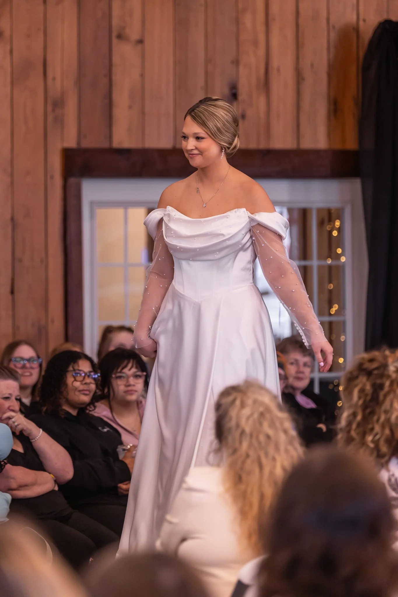 A woman in a white off-shoulder wedding gown walking on runway during a fashion show, audience watching in the background.