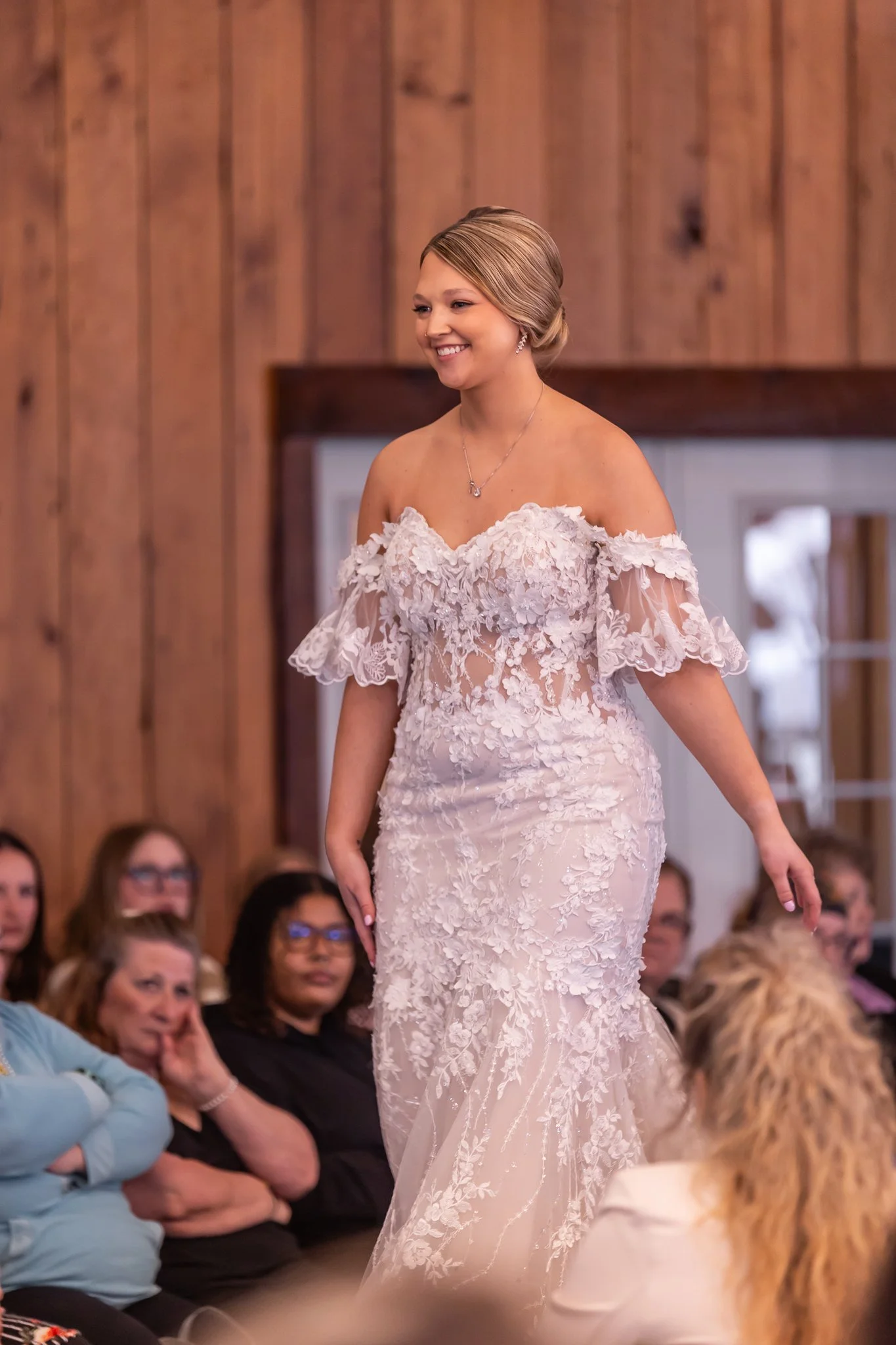 A bride walking down the aisle in a lace wedding dress with off-the-shoulder sleeves, smiling, surrounded by seated guests in a wooden-paneled room.