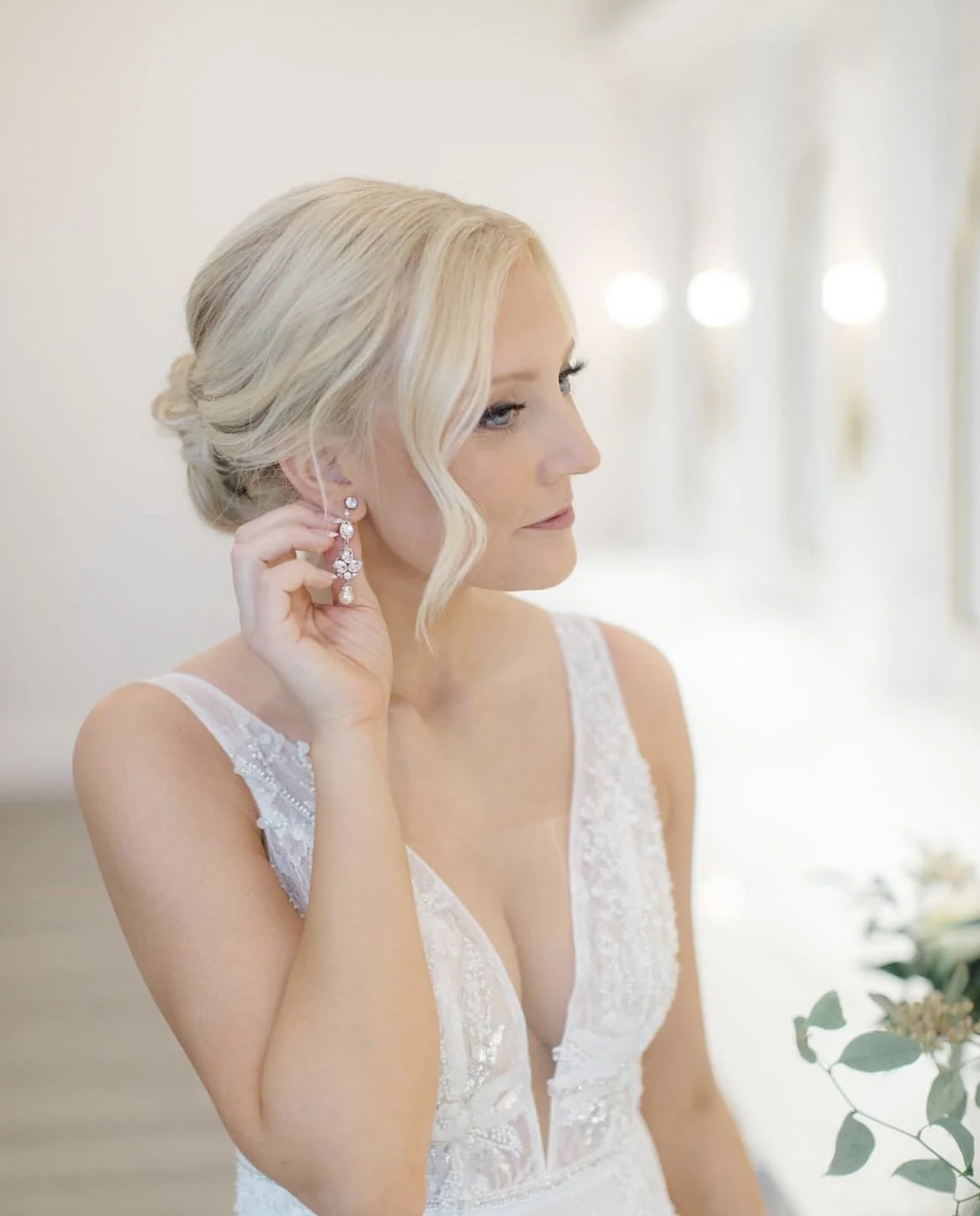 Bride with blonde hair and drop earrings, adjusting her earring in a white lace wedding dress in a bright, elegant room.