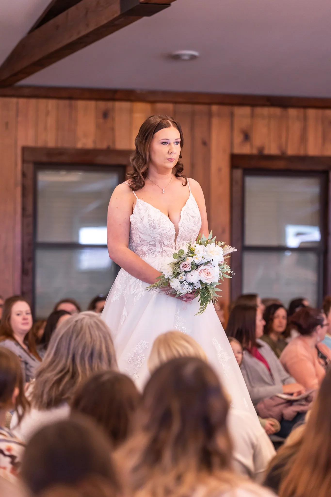 A bride in a white wedding dress holding a bouquet of flowers standing among seated wedding guests in a wood-paneled room.