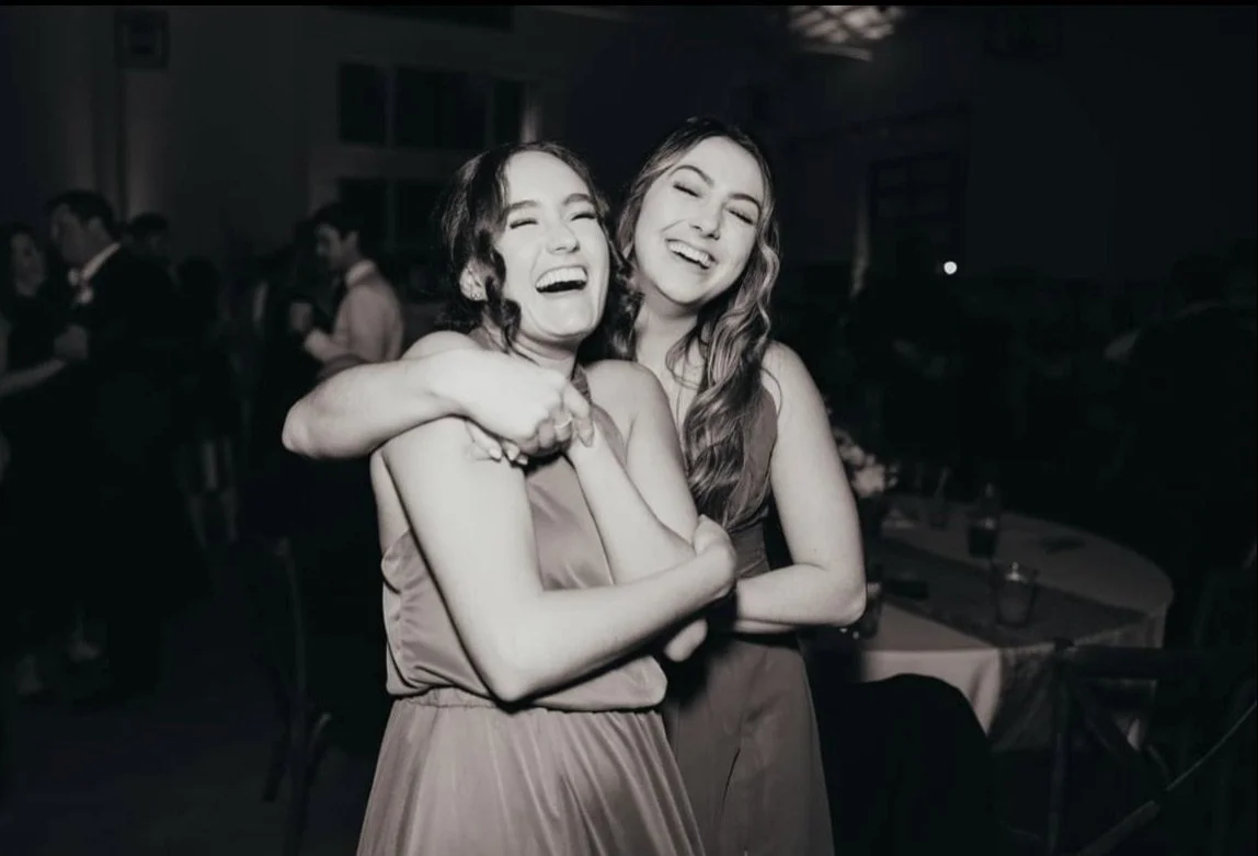 Two women embracing and laughing at a social event or party with other people in the background.