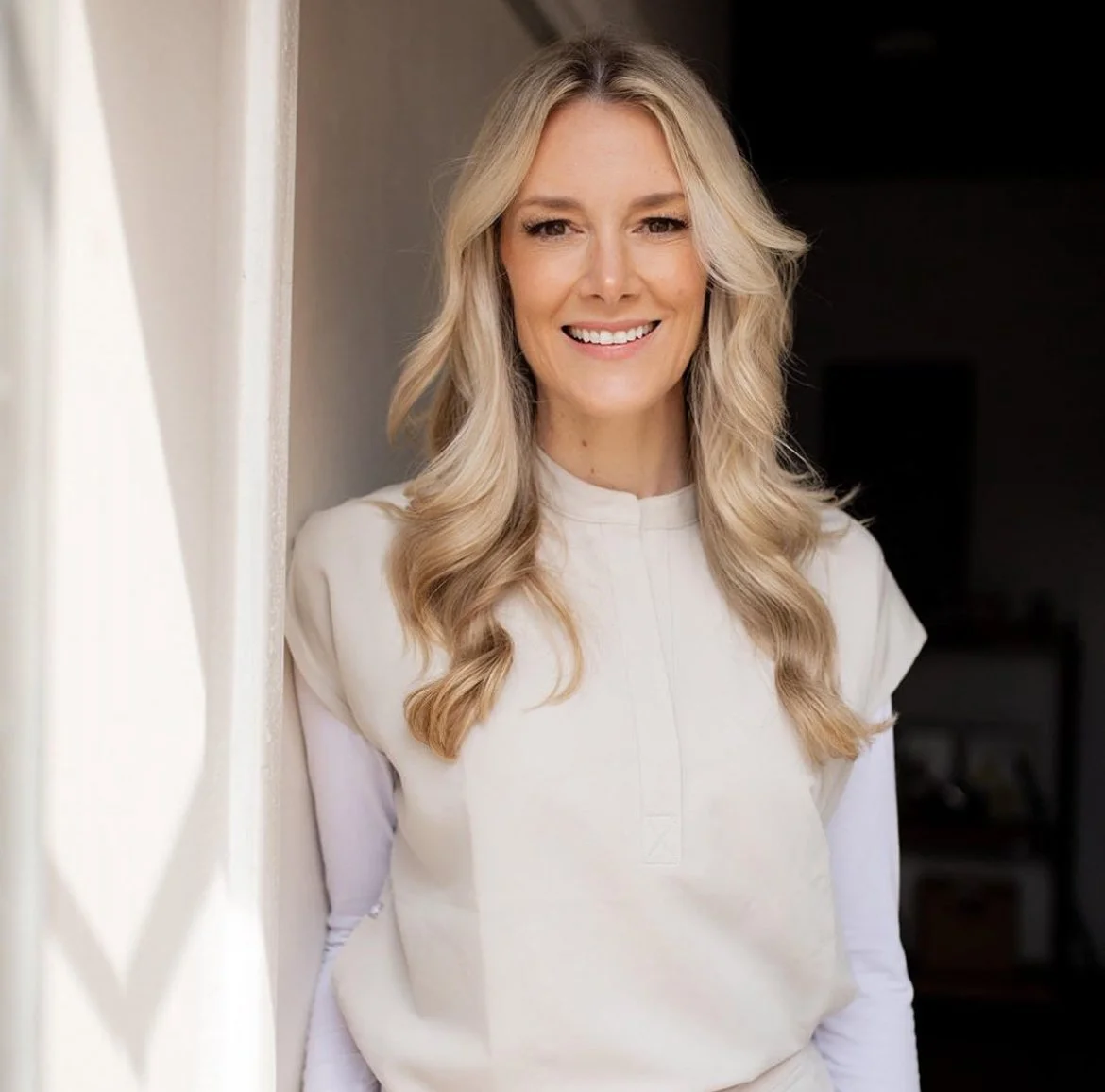 A woman with long blonde wavy hair, smiling, wearing a cream-colored top, standing near a wall in a well-lit room.