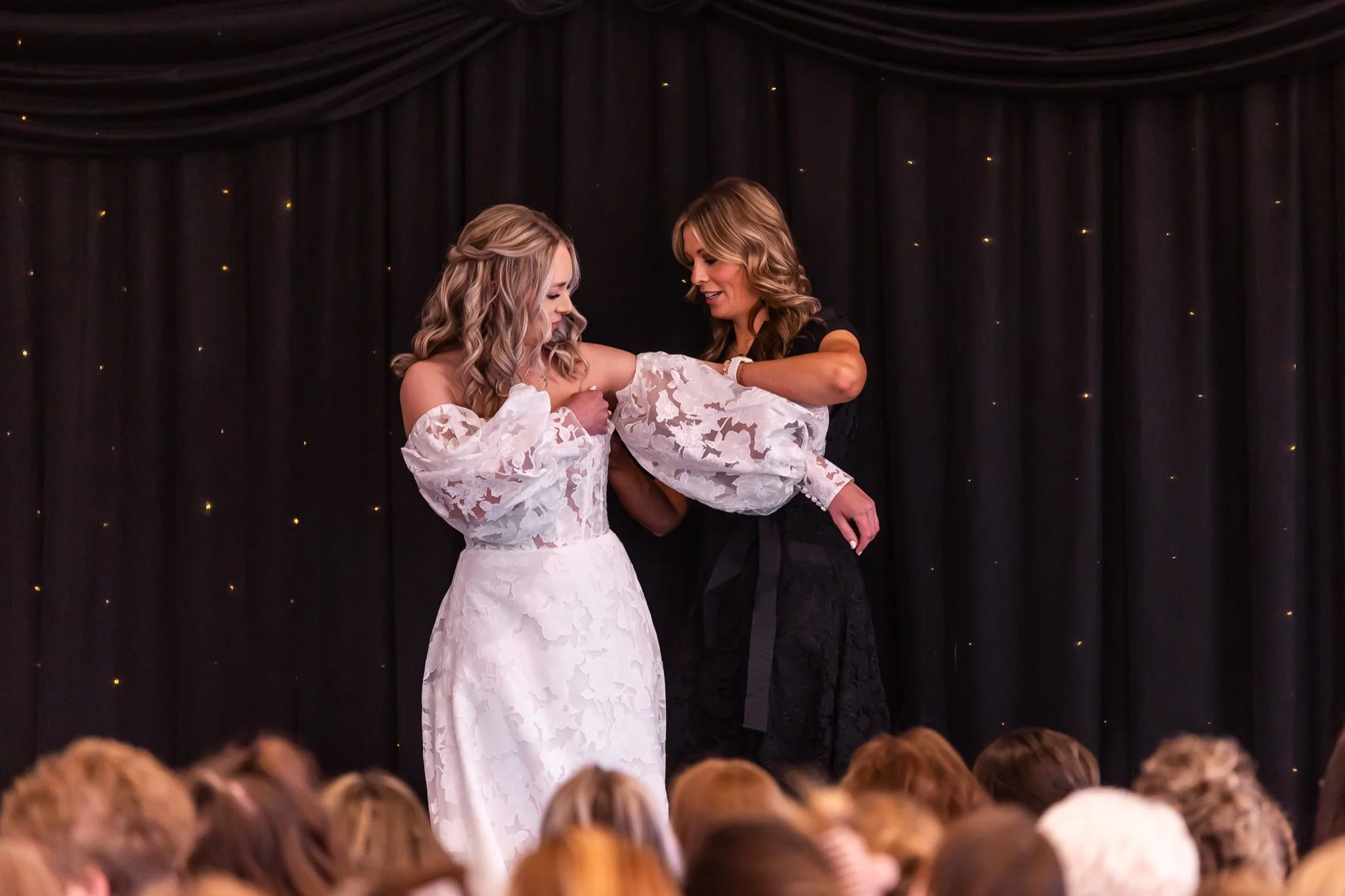 A wedding ceremony where a bride in a white lace dress is receiving her wedding ring from a woman, on a stage with black curtains and small lights, with an audience watching.