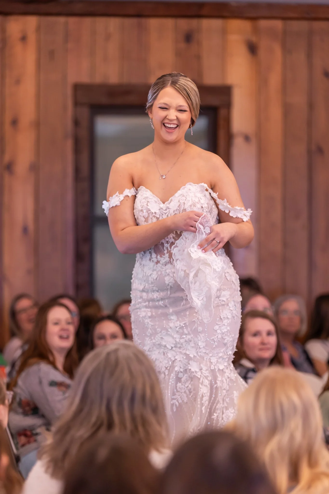 A bride in a white lace wedding gown is smiling and holding a garter in a wooden-paneled room with seated guests watching.