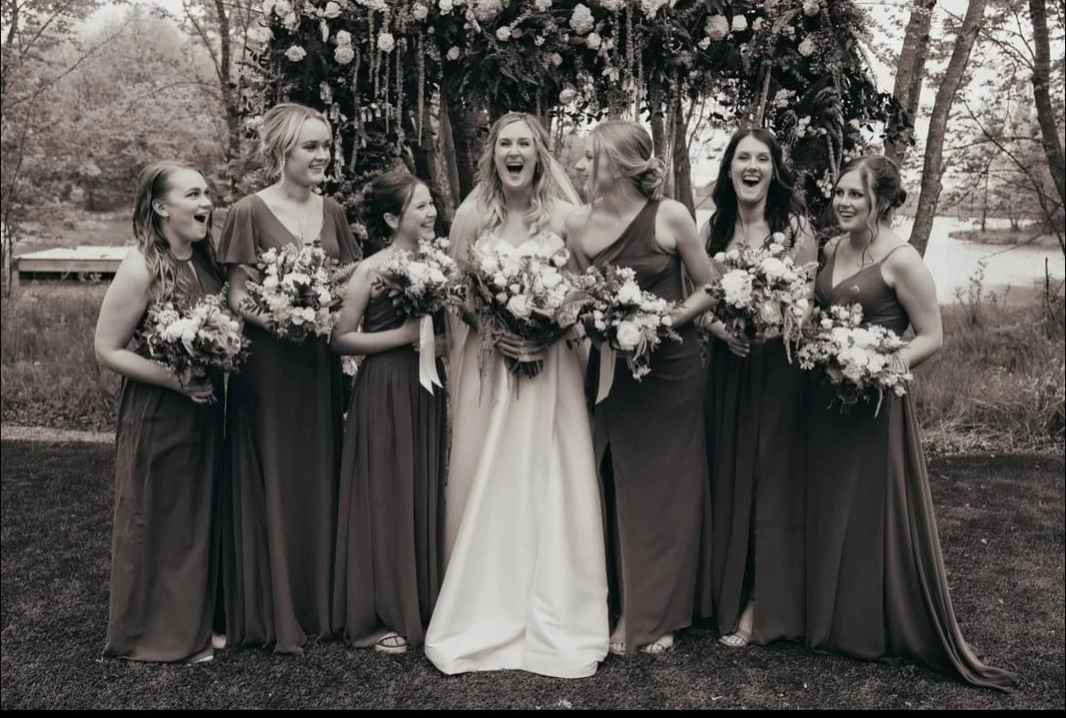 A group of seven women dressed in wedding and bridesmaid dresses, holding bouquets, smiling and laughing outdoors with trees and a body of water in the background.