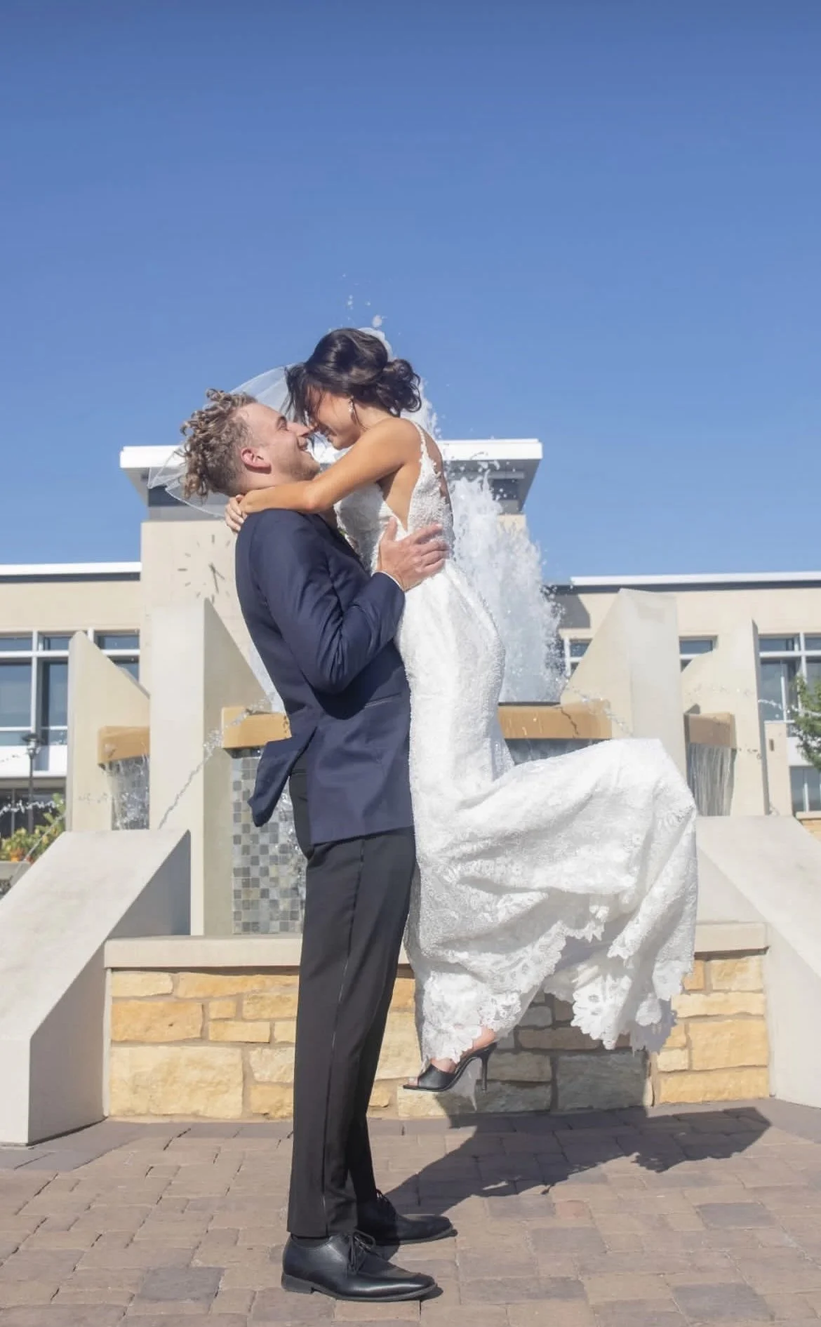 A bride and groom are celebrating on their wedding day in front of a fountain. The groom is lifting the bride, who is wearing a white wedding dress, and they are smiling at each other. The bride is slightly wet from the fountain spray.