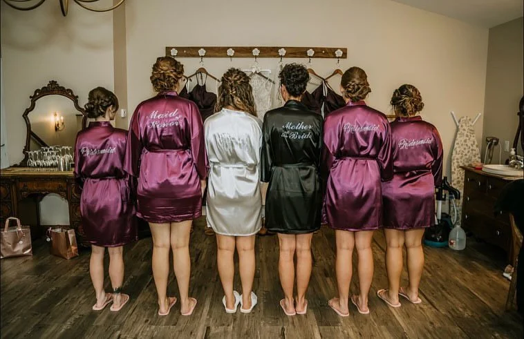 Six women standing with their backs to the camera, wearing satin robes, in a room with wooden floors and a vanity with a mirror.
