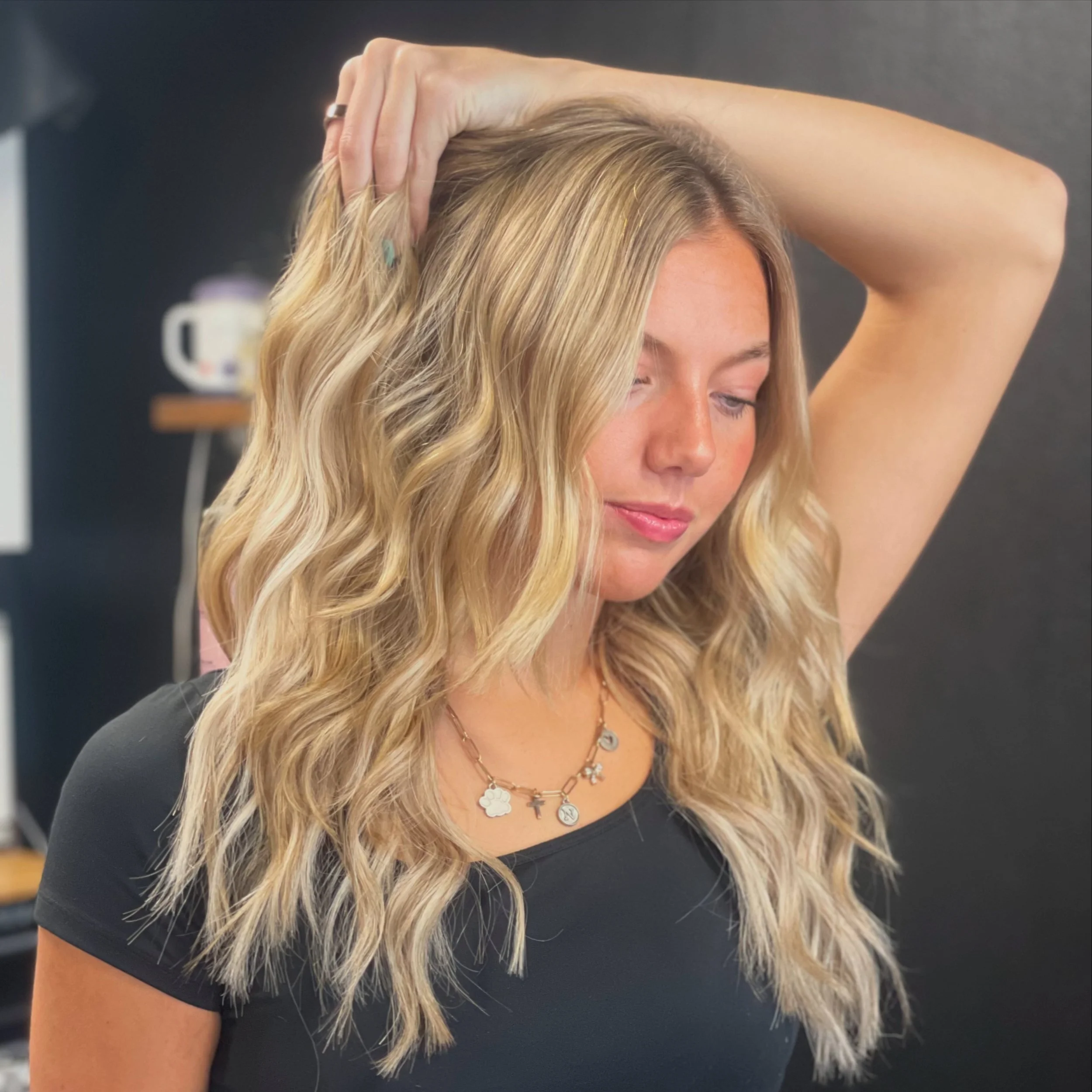 Woman with blonde, wavy hair, wearing a black top and layered necklaces, with her hand on her head, indoors against a dark background.