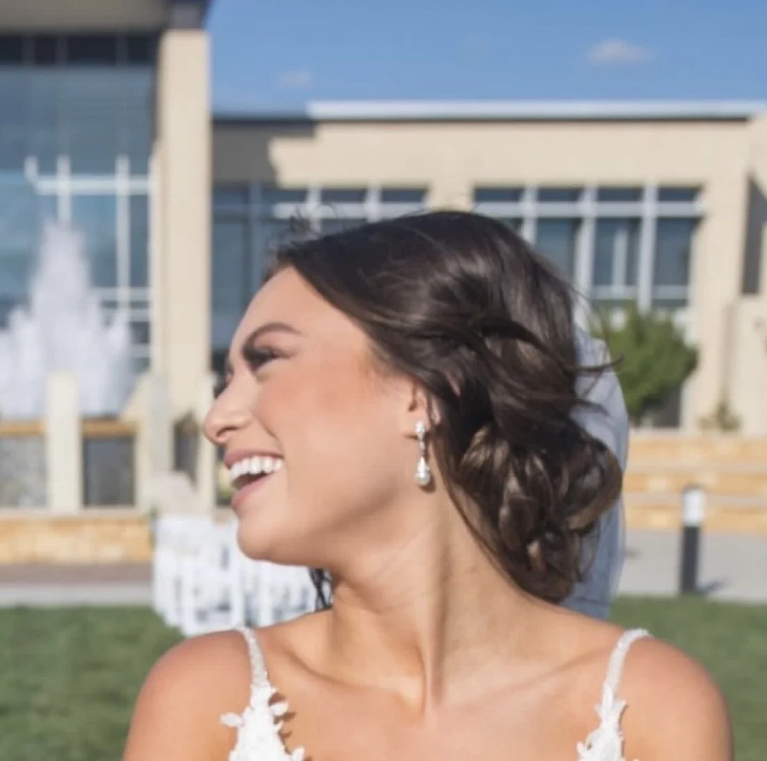A woman smiling and looking to the side outdoors near a modern building with a fountain in the background.
