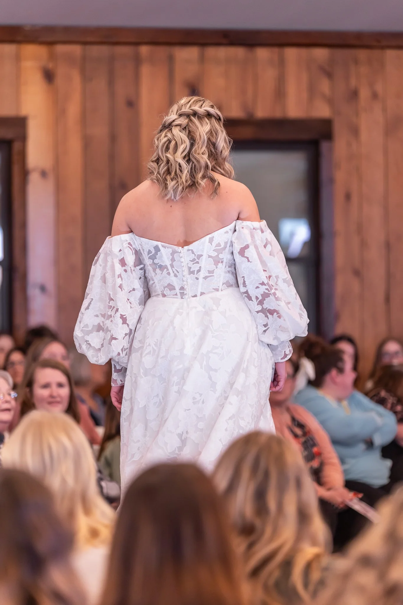 Woman in white lace off-shoulder dress walking in a room with wooden walls, with women seated and looking on.